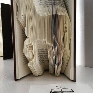 May include: A book art sculpture of a horse, folded from the pages of a book. The book is bound in brown leather and the pages are cream colored. The sculpture is displayed on a white surface with a business card that reads "THE BOOK MAGICIAN".