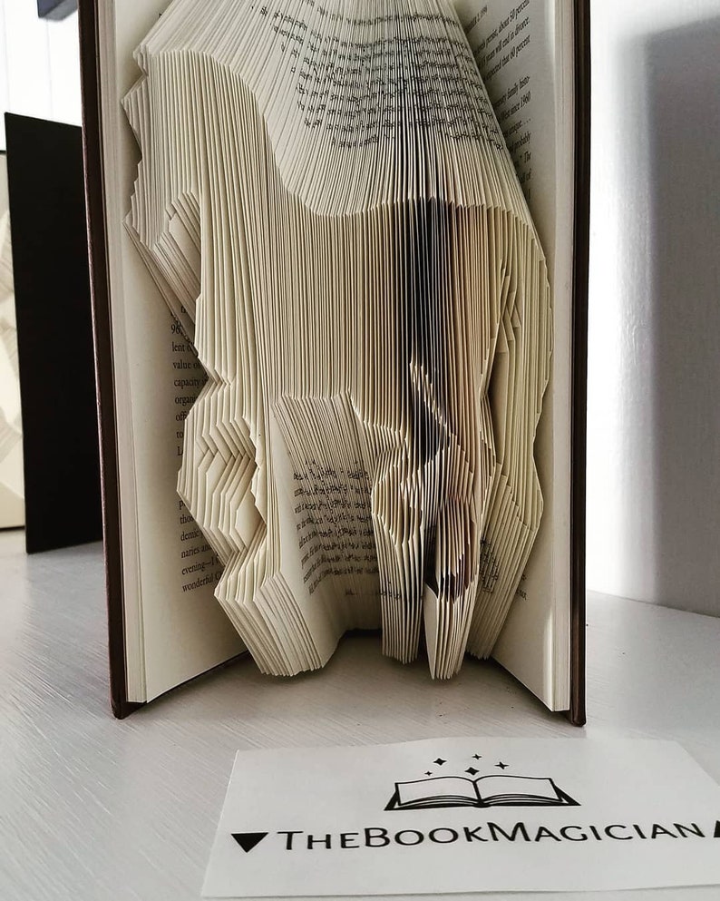May include: A book art sculpture of a horse, folded from the pages of a book. The book is bound in brown leather and the pages are cream colored. The sculpture is displayed on a white surface with a business card that reads "THE BOOK MAGICIAN".