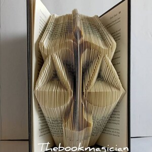 May include: A hardback book with pages folded to create a three-dimensional sculpture of a scale of justice. The book is open, displaying printed text on the pages. The white text "Thebookmagician" is visible at the bottom of the image.