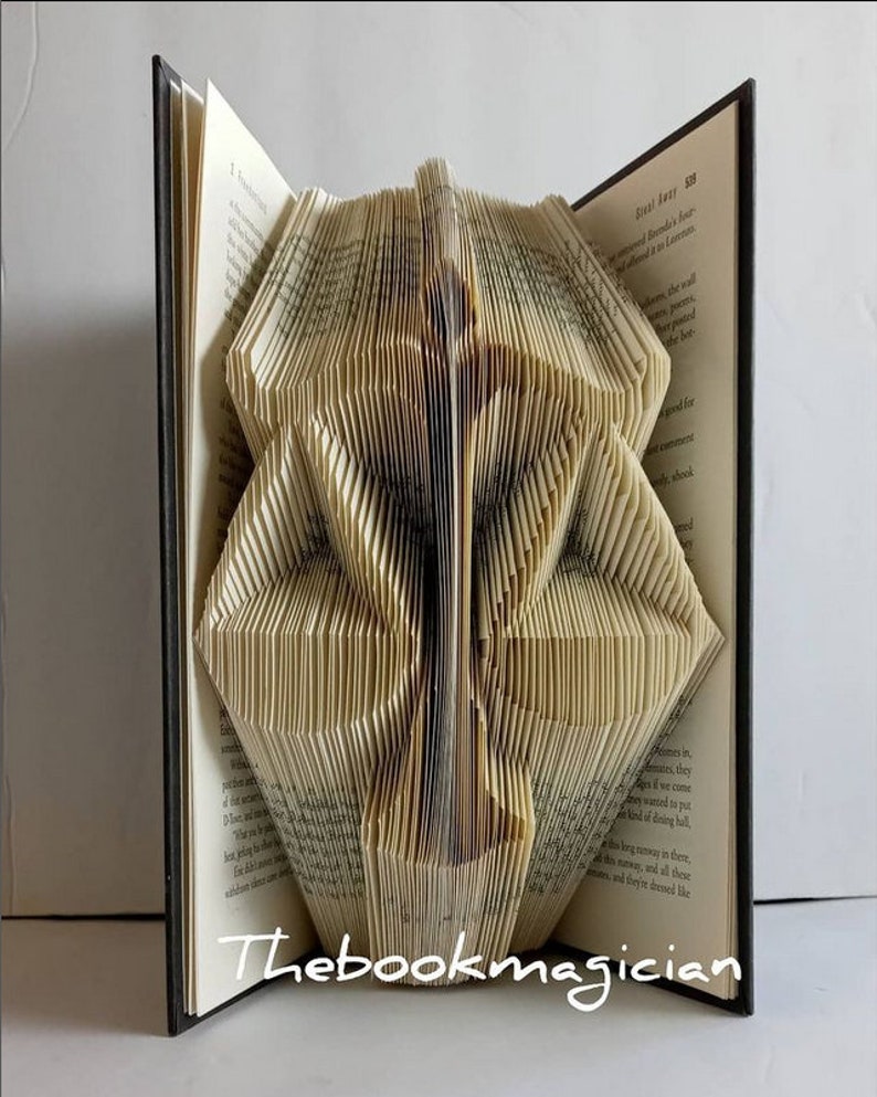 May include: A hardback book with pages folded to create a three-dimensional sculpture of a scale of justice. The book is open, displaying printed text on the pages. The white text "Thebookmagician" is visible at the bottom of the image.