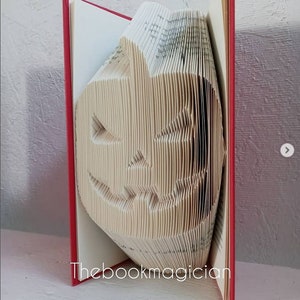 May include: A book folded into the shape of a jack-o'-lantern. The book has a red cover and the pages are folded to create a 3D effect. The book is titled "Thebookmagician".