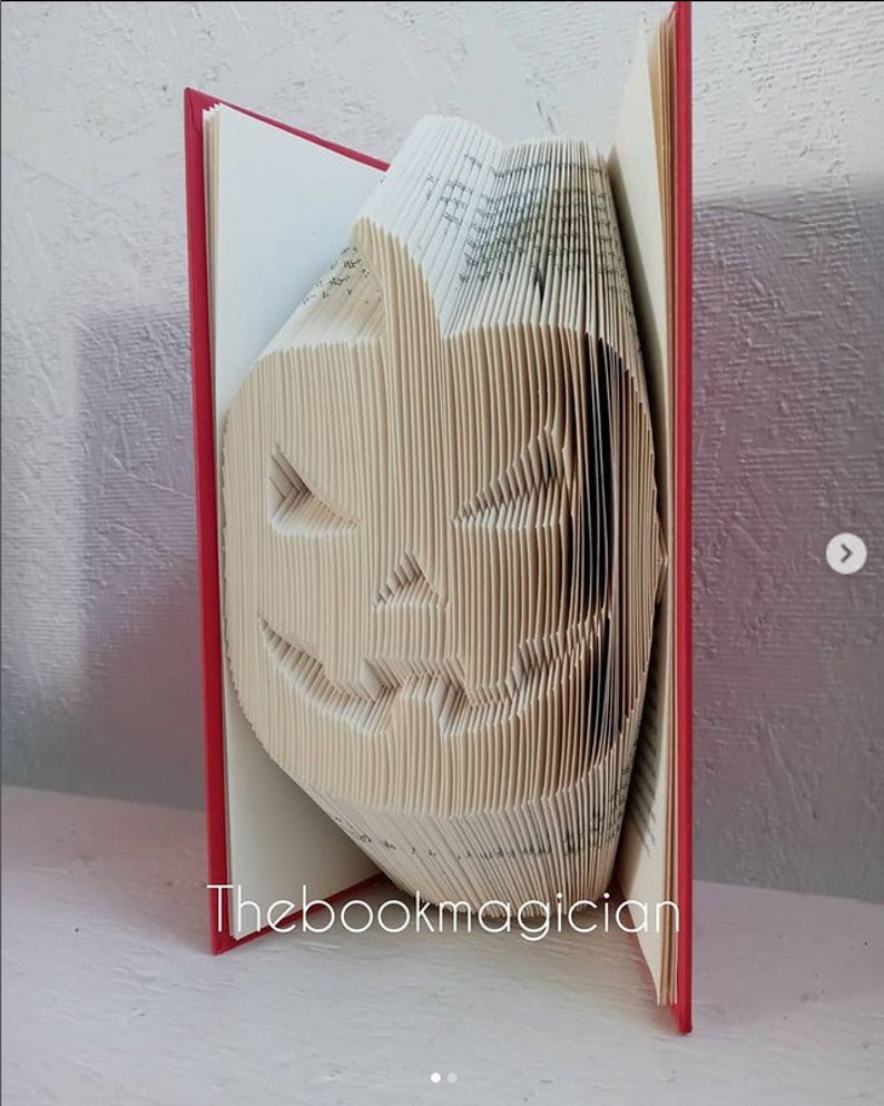 May include: A book folded into the shape of a jack-o'-lantern. The book has a red cover and the pages are folded to create a 3D effect. The book is titled "Thebookmagician".