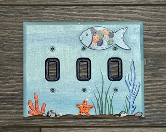 May include: A light blue triple light switch cover with a painted ocean scene. The scene includes a fish with orange and green spots, a starfish, seaweed, and coral.