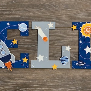 May include: Wooden letters spelling "ELI" painted in a space theme. The letters are blue with white stars, planets, and a rocket ship. The letter "I" is gray with a comet and stars.