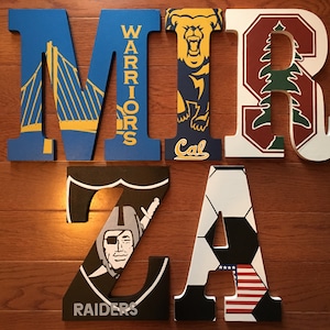 May include: Wooden letters spelling out the name "MIRZA" with different sports team logos and designs. The "M" is blue with a Golden Gate Bridge design and "WARRIORS" written vertically on the side. The "I" is blue with a California Golden Bears logo and "Cal" written below. The "R" is maroon with a Stanford University logo. The "Z" is black with a Las Vegas Raiders logo and "RAIDERS" written below. The "A" is white with a soccer ball design and an American flag.