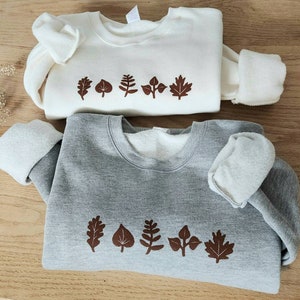 May include: Two sweatshirts with a leaf design. The top sweatshirt is white with brown leaves. The bottom sweatshirt is gray with brown leaves.