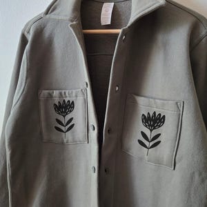 May include: A sage green button-up shirt with two front pockets, each featuring a black floral design. The shirt has a collared neckline and is hanging on a wooden hanger.