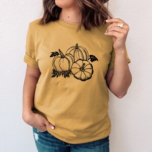 Peut inclure: Un t-shirt jaune moutarde avec un motif noir et blanc de trois citrouilles.
