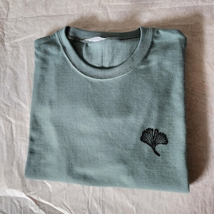 Puede incluir: Una camiseta verde salvia doblada, con cuello redondo y un diseño de hoja de ginkgo negro bordado en la parte inferior izquierda. La camiseta es de material suave y está sobre una superficie texturizada blanquecina.