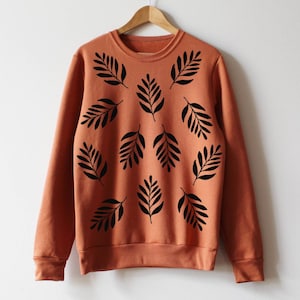 May include: A terracotta-coloured sweatshirt with a crew neck and long sleeves. The front displays a pattern of black leaf designs. The sweatshirt is suspended on a wooden hanger against a white backdrop.