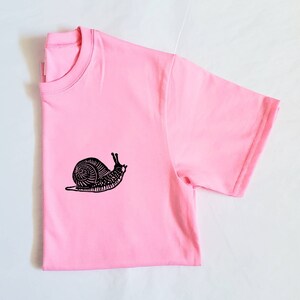 May include: A pink t-shirt with a black snail graphic printed on the front.