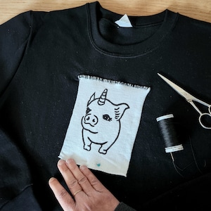 Op de afbeelding: Zwart sweatshirt met een witte patch met een cartoonvarken met een eenhoornhoorn. De patch is op het sweatshirt gespeld. Schaar en een spoel zwart garen liggen ernaast.