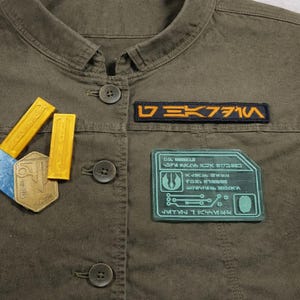 May include: Olive green jacket with several patches and pins. The jacket has a button-down front and a collar. Patches include a rectangular patch with orange text, a teal patch with white text, and several gold-colored pins.