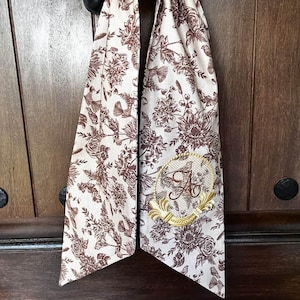 May include: A cream-colored fabric wreath bow with a brown floral pattern and a gold monogram "A" in a circle. The bow is tied around a wooden door.