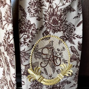 May include: A beige fabric scarf with a brown floral pattern. The scarf has a gold embroidered monogram with the letter "A" in a decorative frame.