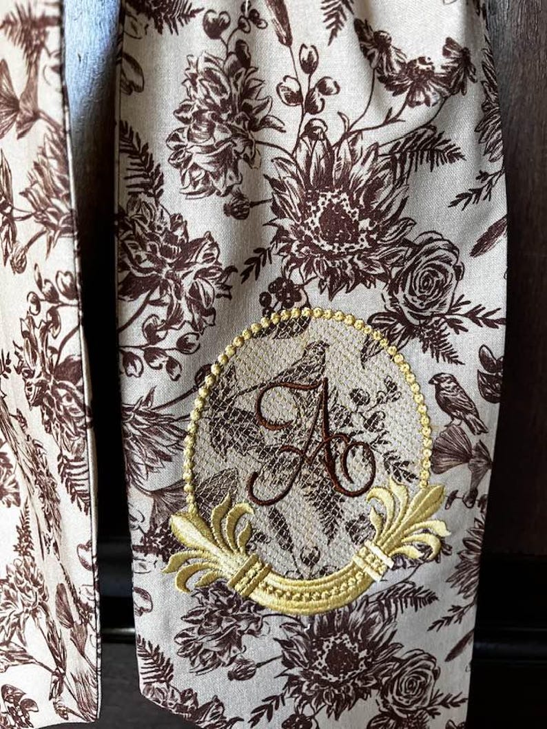 May include: A beige fabric scarf with a brown floral pattern. The scarf has a gold embroidered monogram with the letter "A" in a decorative frame.