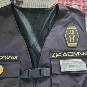 May include: A black vest with gold embroidered patches. The patches include the text "71WV", "MAZD", "AZW", "AKAOV", "NAME: KYLE MAKWELL", "SUPER THE BACC LEAVES", and "LEVEL 7 ACCCES".