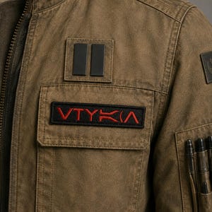 May include: A brown jacket featuring a stand-up collar, a front chest pocket with a black rectangular patch displaying the red text VTYKA, a second black patch with two vertical bars, and a sleeve pocket containing three dark pens.