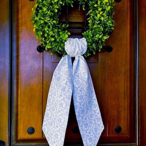 May include: A green boxwood wreath with a white and light blue patterned ribbon hanging on a dark brown wooden door. The wreath is round and full, with a classic design. The ribbon is tied in a bow.