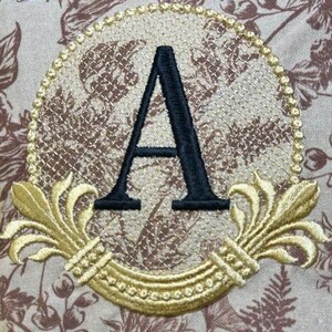 May include: A gold embroidered monogram with the letter "A" in black, surrounded by a decorative floral design.