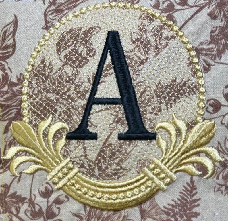 May include: A gold embroidered monogram with the letter "A" in black, surrounded by a decorative floral design.