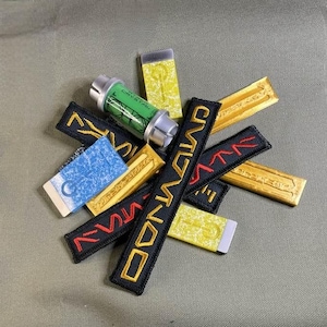 May include: A collection of fabric patches with various designs and colors, including black, gold, red, and green. The patches feature text such as "COMMAND" and "ZYNA", and some have intricate patterns. There is also a green and white cylindrical object with the text "CRYSTAL" on it.