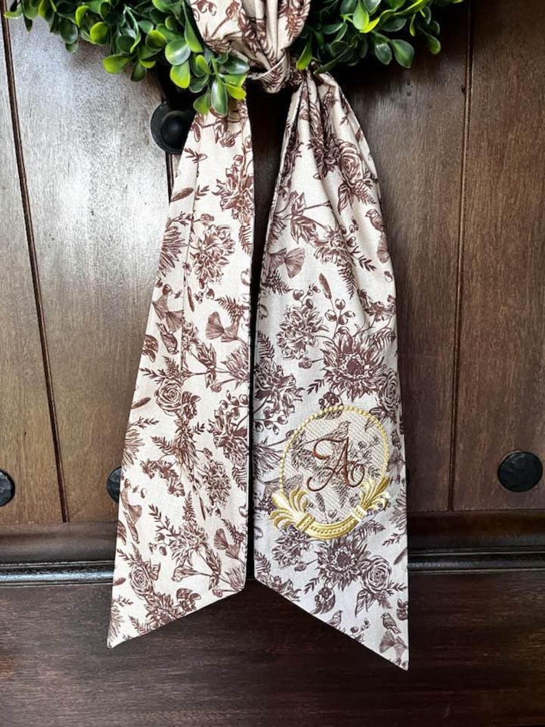 May include: A cream-colored fabric wreath bow with a brown floral pattern and a gold monogram "A" in a circle. The bow is tied around a wooden door.