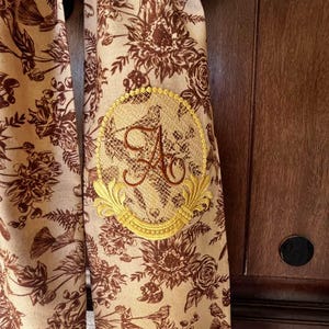 May include: A beige fabric scarf with a brown floral print and a gold embroidered monogram of the letter "A" in a circle.