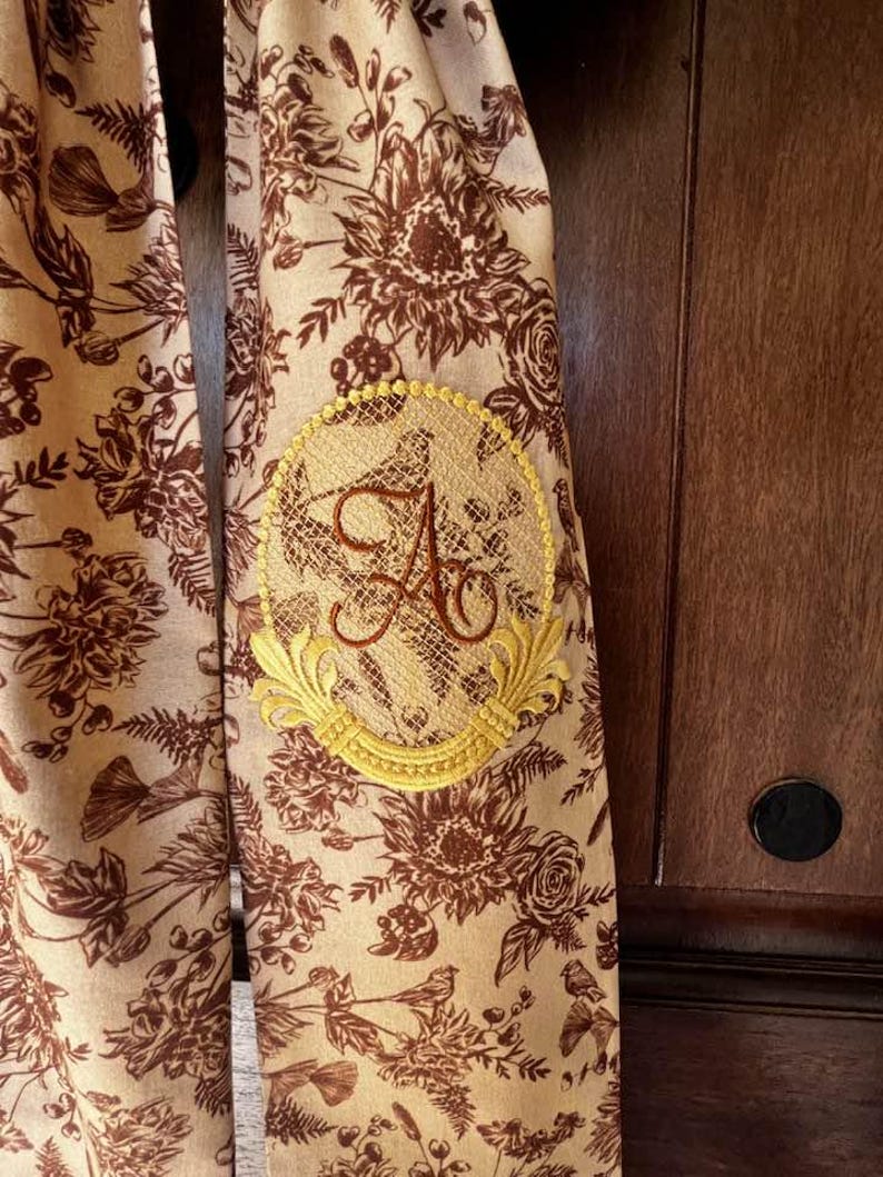 May include: A beige fabric scarf with a brown floral print and a gold embroidered monogram of the letter "A" in a circle.