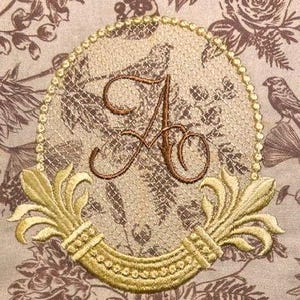 May include: Embroidered monogram with a gold frame and floral accents. The letter "A" is in a script font and is surrounded by a delicate floral pattern. The frame is adorned with a beaded border and two stylized floral motifs.