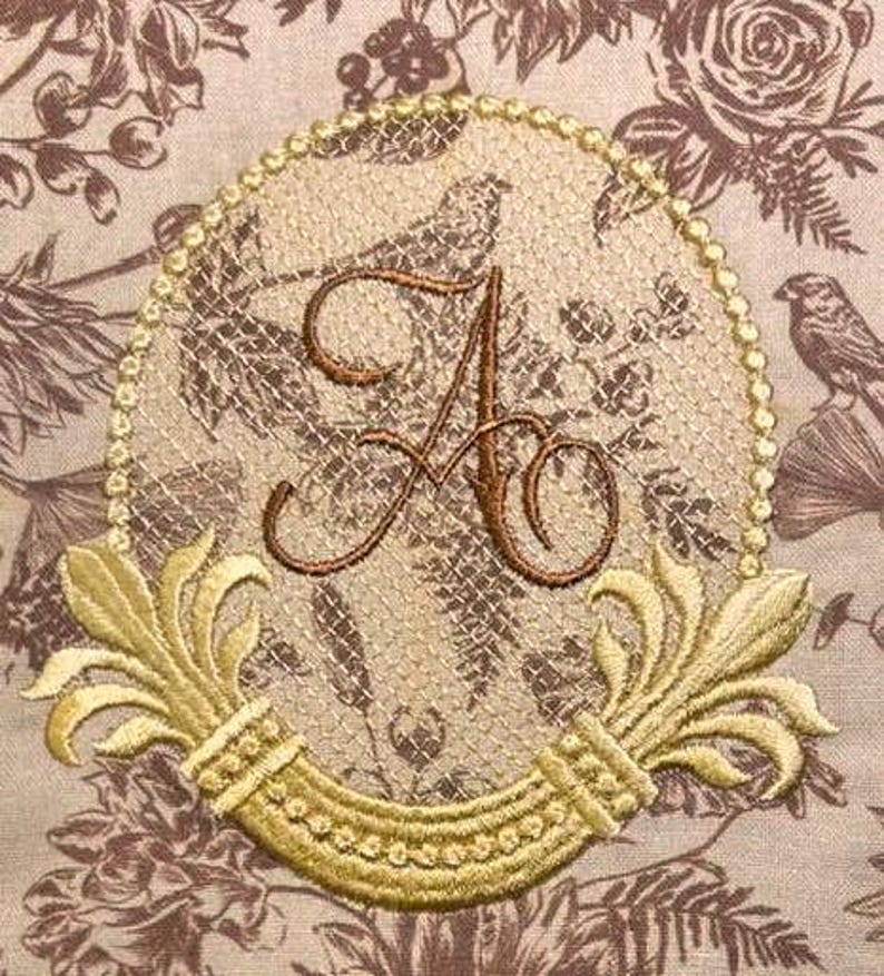 May include: Embroidered monogram with a gold frame and floral accents. The letter "A" is in a script font and is surrounded by a delicate floral pattern. The frame is adorned with a beaded border and two stylized floral motifs.