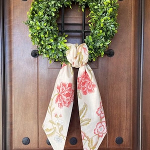 May include: A green boxwood wreath with a floral patterned fabric bow. The bow is tied in a knot and the ends are hanging down. The wreath is hanging on a wooden door.