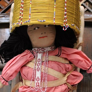 May include: A handmade doll with a pink dress, brown boots, and a yellow woven hat with beaded trim. The doll is wearing a beaded necklace and has a white face with painted features.