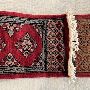 May include: A red runner rug with a geometric pattern and white fringe. The rug has a traditional design with intricate details.