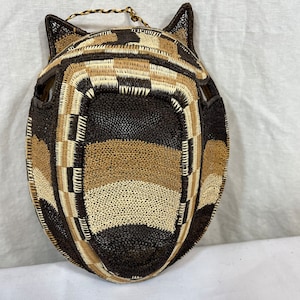 May include: A woven mask with a brown, tan, and white pattern. The mask has a rounded shape with two small ears on top.