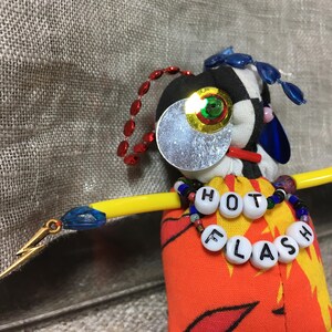 May include: A handmade doll with a black and white checkered head, a large silver eye, and a yellow arm. The doll's torso is orange with a flame design. A necklace spells out "HOT FLASH".