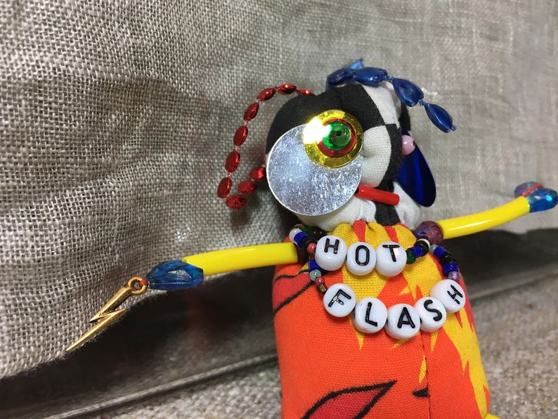 May include: A handmade doll with a black and white checkered head, a large silver eye, and a yellow arm. The doll's torso is orange with a flame design. A necklace spells out "HOT FLASH".