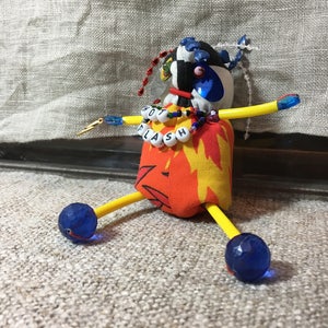 May include: A handmade doll with a fabric body featuring a flame design. The doll has yellow arms and legs, blue faceted bead feet, and a head with a checkered pattern. The doll's necklace spells "HOT FLASH".