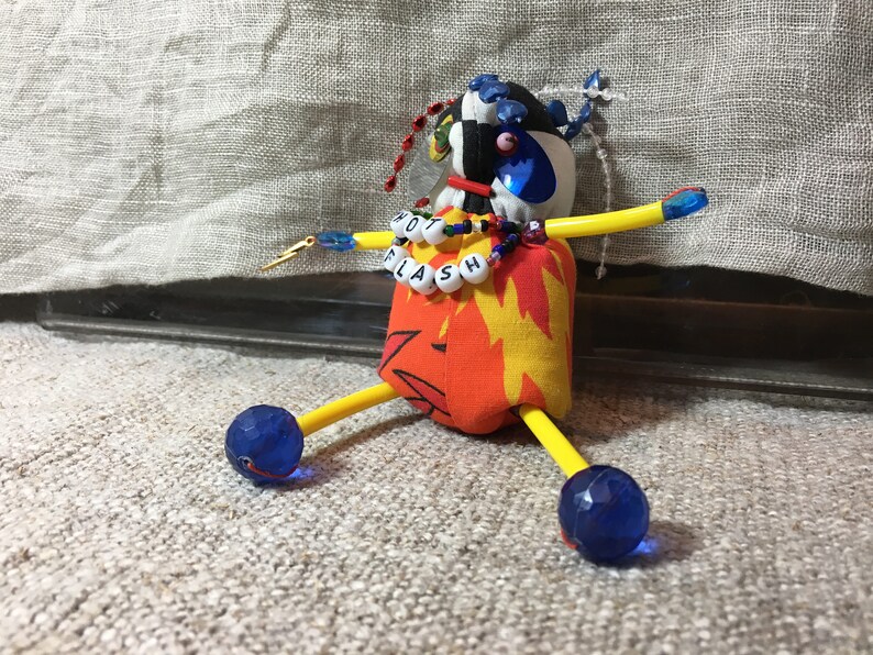 May include: A handmade doll with a fabric body featuring a flame design. The doll has yellow arms and legs, blue faceted bead feet, and a head with a checkered pattern. The doll's necklace spells "HOT FLASH".