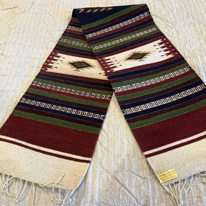 May include: A woven table runner with a geometric pattern in shades of red, green, white, and blue. The runner has fringe on the ends and a small tag that reads "El Segundo Leather".