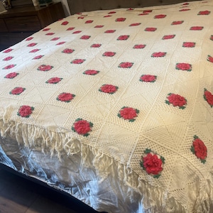 May include: Cream-colored, handmade crochet blanket with a repeating pattern of red roses and green leaves. The blanket has a fringed edge and is displayed on a bed. A vintage, floral-themed home decor item.