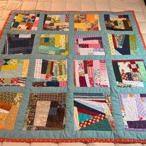 May include: A handmade quilt with a patchwork design. The quilt features a light blue border and a red polka-dot trim. The patchwork squares are filled with various fabric patterns and colors, including floral, striped, and solid fabrics.
