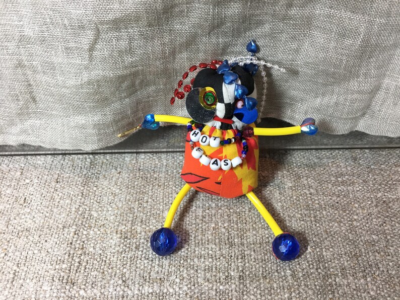 May include: A whimsical handmade doll with a black and white head, blue and red accents, and yellow arms and legs. The doll's torso is orange with a floral pattern and the text "HOT PEAS". Blue gem-like spheres form the feet.