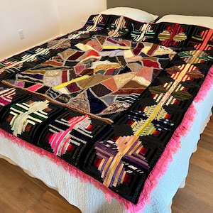 May include: A large patchwork quilt with a black background and a variety of colorful fabrics. The quilt is made up of squares and rectangles of different sizes and colors, and it has a pink fringe around the edges.
