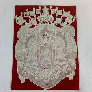 May include: White lacework depicting a coat of arms with two lions, a crown, and flags. The text "Gott macht das" is visible at the bottom.