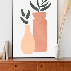 May include: A framed print of a minimalist illustration of two vases with green leaves. The vases are peach and terracotta colored and are on a white background.