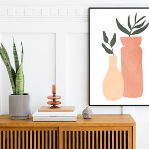 May include: A framed print with a minimalist design featuring two abstract vases with green leaves in a peach and terracotta color scheme. The print is hanging on a white wall above a wooden cabinet with a potted plant.