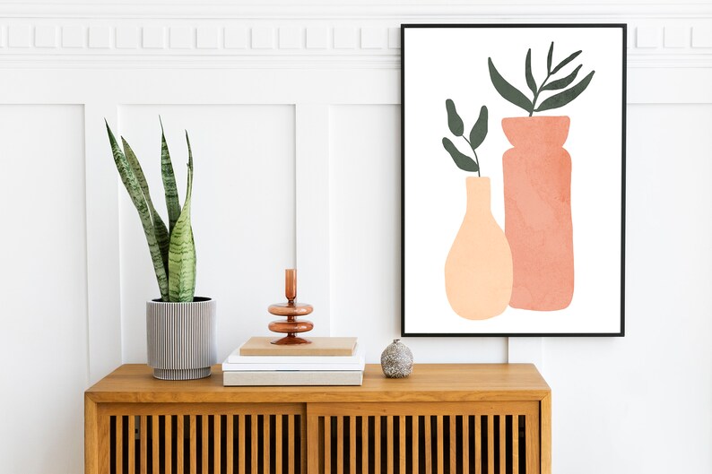 May include: A framed print with a minimalist design featuring two abstract vases with green leaves in a peach and terracotta color scheme. The print is hanging on a white wall above a wooden cabinet with a potted plant.