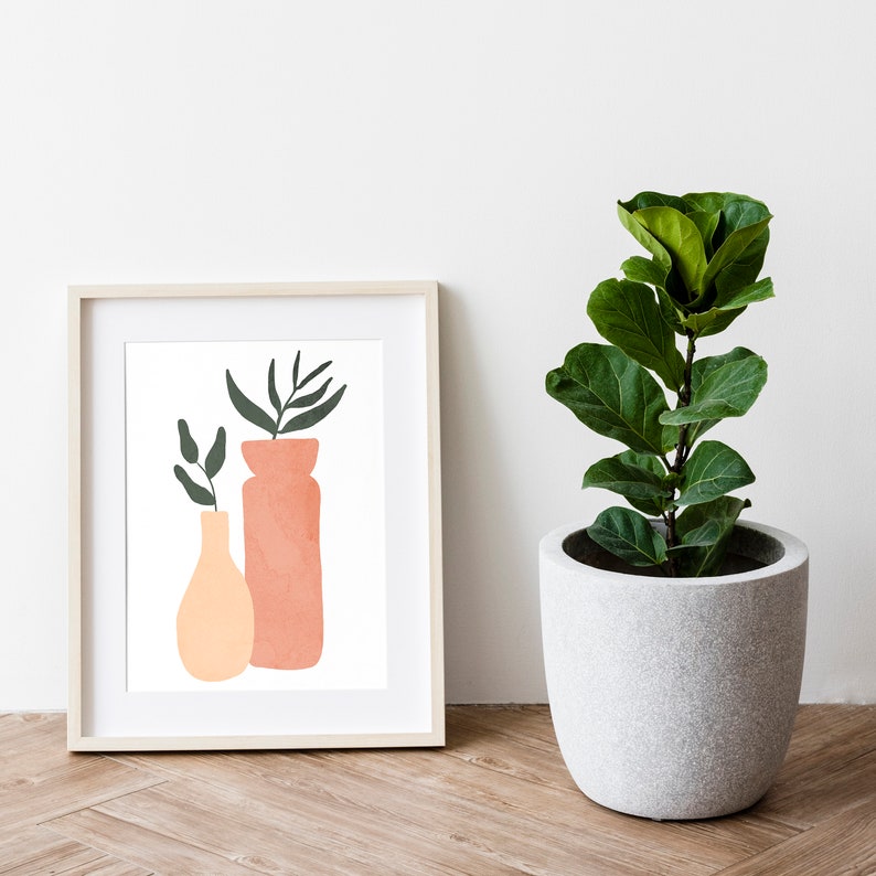 May include: A framed print of a minimalist illustration of two vases with green leaves. The vase on the left is peach and the vase on the right is pink. The print is on a white background and is framed in a light wood frame.
