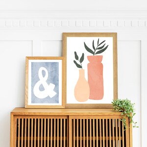May include: Two framed prints on a wooden cabinet. The print on the left has a blue background with a white ampersand symbol. The print on the right has a white background with a green plant and two orange and pink vases.